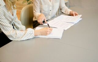 woman signing on white printer paper beside woman about to touch the documents